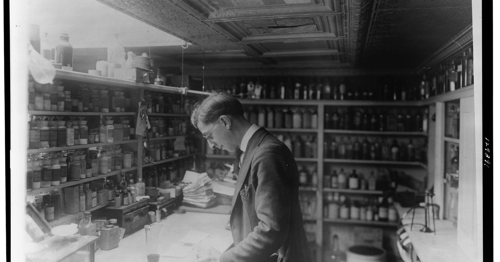 Archiwalne czarno-białe zdjęcie farmaceuty przy ladzie apteki wśród półek pełnych aptecznych butelek