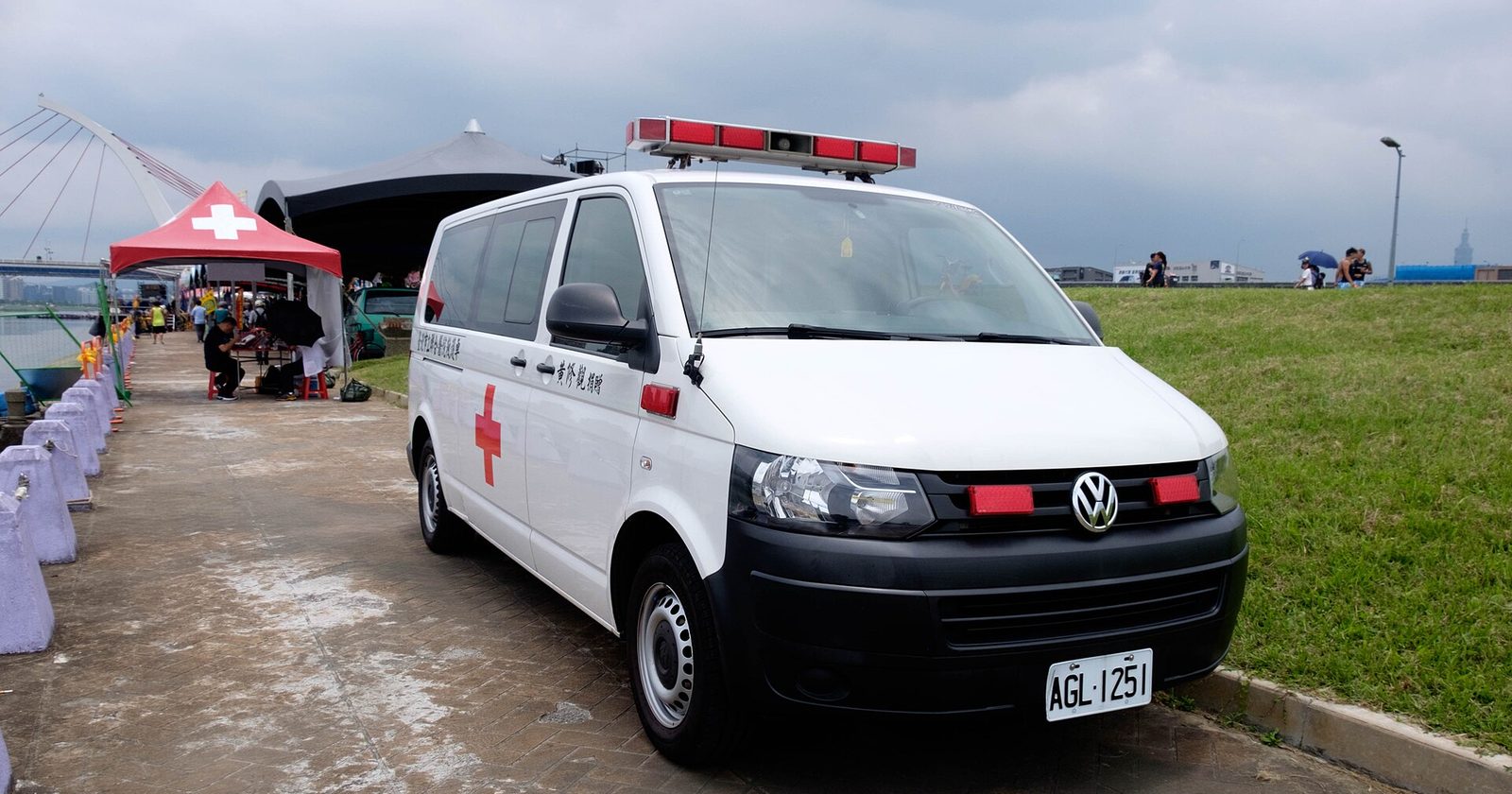 Biały ambulans z czerwonym krzyżem zaparkowany obok czerwonego namiotu pierwszej pomocy nad rzeką